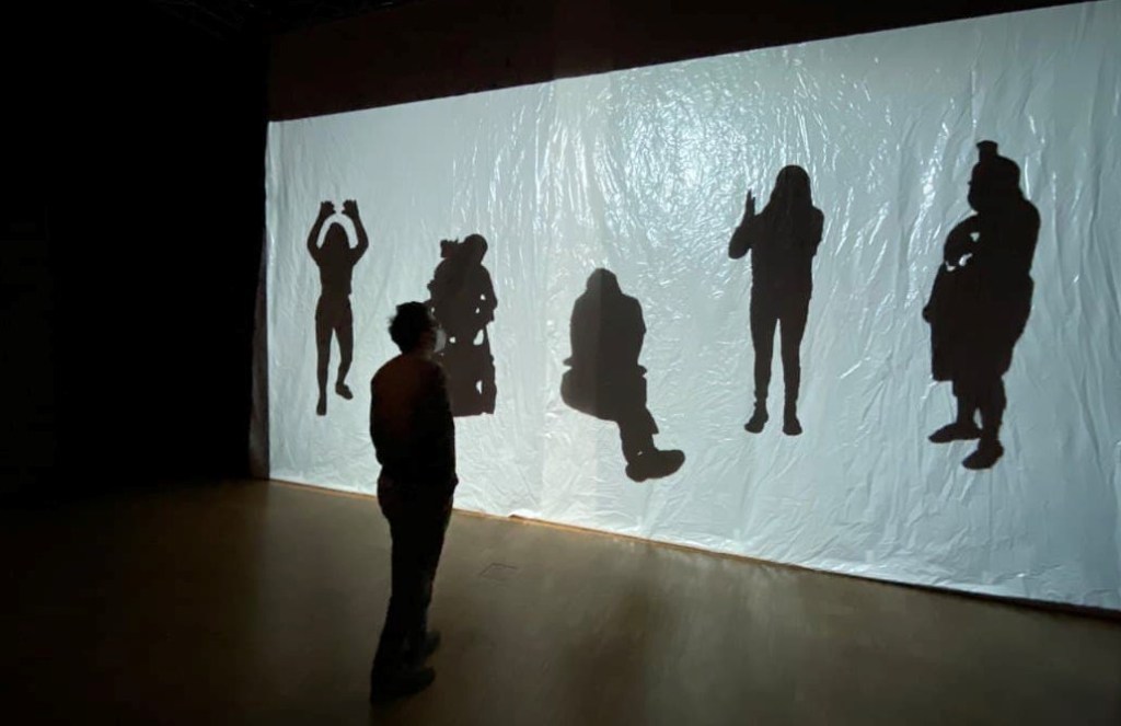 Zu sehen ist ein Bild der Installationssituation: Vor einer großen Leinwand steht ein Besucher. Auf der Leinwand sind fünf lebensgroße Silhouetten zu sehen.