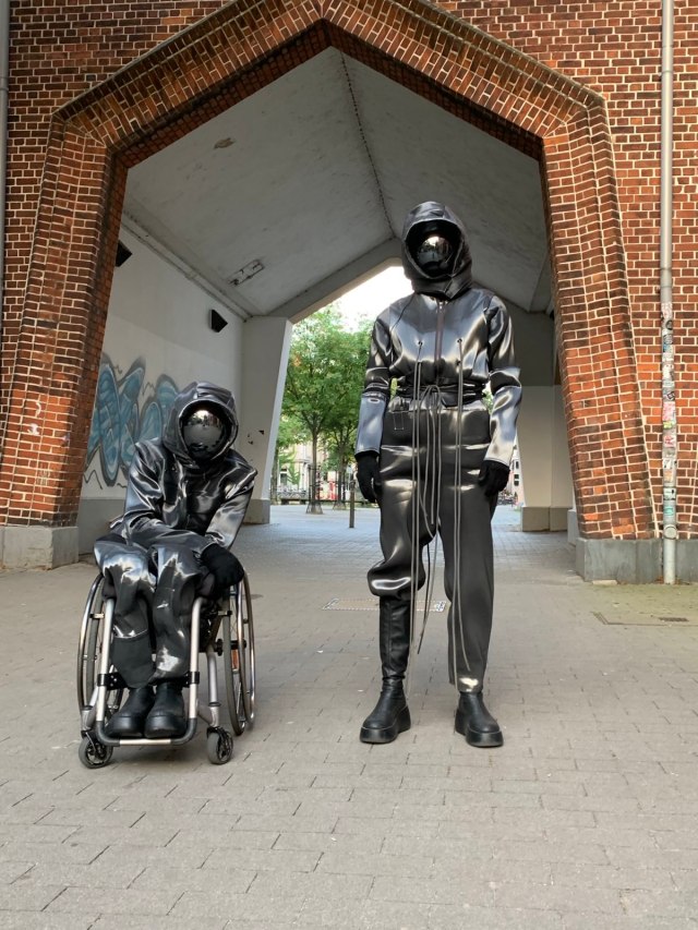 Vor ein Durchgang, der auf einen Platz führt stehen zwei Astronauten*innen. Sie tragen silbern schimmernde Anzüge und spiegelnde Visiere. Einer der beiden Astronautinnen sitzt im Rollstuhl. Die andere steht. Beide schauen sie in die Kamera.